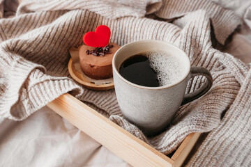 ceramic grey cup with black coffee and round small chocolate cheesecake with red heart in cozy atmosphere on crumpled fabric of bed. Surprise for loved , gift idea for Valentine's Day, selective focus