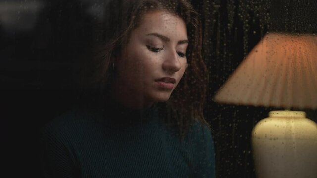 Pretty woman looking out through her home window on a rainy nights a water drips down. Cinematic