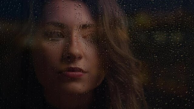 Close up of woman looking at camera with hand on window on rainy night. Drops of water on window are in focus. Cinematic