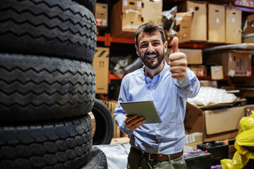 Smiling bearded excited businessman standing in storage of export firm, holding tablet and showing...
