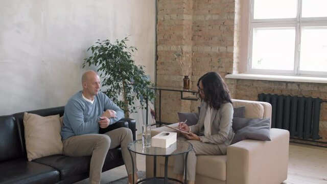 Tilt down of professional female therapist writing notes on clipboard and talking to emotional middle-aged man sitting on couch in private practice office