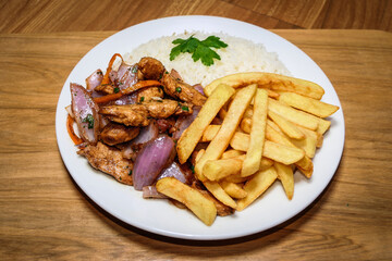 Peruvian food saltado chicken, french fries, seasoned chicken, onions, tomatoes, roasted vegetables, white arros on a white plate.