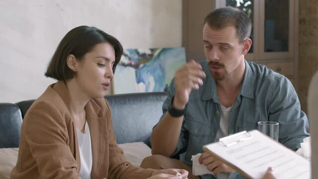 Medium shot of young couple sharing their problems with unrecognizable therapist with clipboard during session in private practice