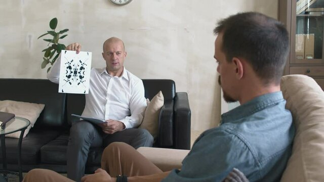 Tracking shot of middle-aged male psychotherapist performing Rorschach test on young bearded man sitting in chair during session
