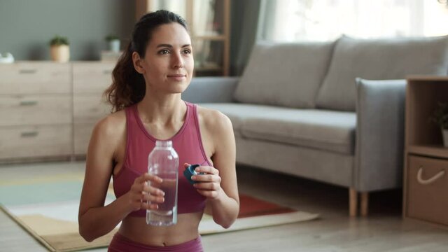 Waist-up shot of brown-eyed sporty Caucasian young woman with ponytail hairstyle wearing crop top, sitting in living room, drinking water from bottle and smiling