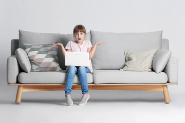 Surprised little girl with laptop on sofa against light background