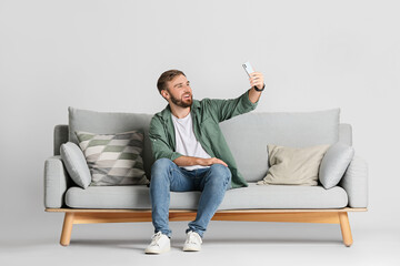 Young man with mobile phone on sofa against light background