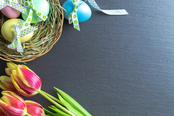 Basket easter decoration: Colourful egg with tape ribbon, spring tulips on dark rough stone background. Congratulatory easter design. Top view.
