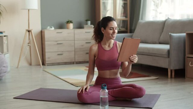 Full Steadicam of attractive fit young Caucasian woman wearing sporty outfit, sitting in lotus pose on yoga mat at home, video calling via tablet computer, waving hand hello, smiling, talking