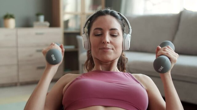 Chest-up POV of young fit Caucasian woman wearing sports top and wireless headphones, leaning on fitball and doing biceps exercises with dumbbells at home, looking and smiling on camera