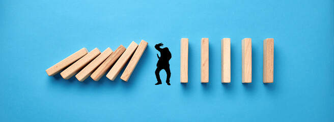 Silhouette of a man in panic try to protect himself against collapsing wooden dominos on blue...