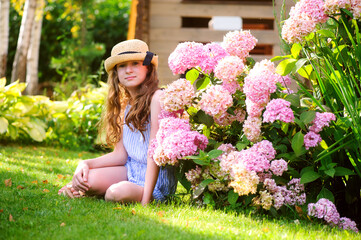 happy kid girl walking in summer garden in straw, posing near pink hydrangea macrofila bush