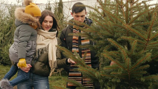Family, winter holidays and people concept - happy mother, father and little daughter choosing christmas tree at street market.
