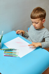 Cute little boy with blond hair draws colored pencils at home. Draws at the blue table.
