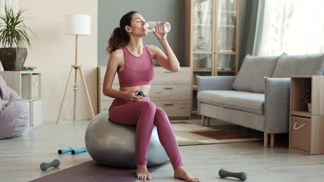 Full shot of fit joyful Caucasian woman wearing yoga outfit sitting on fitball in middle of living room, drinking water from sports bottle and smiling