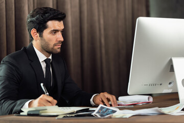 Portrait of young handsome confident businessman in suit working with desktop computer at office