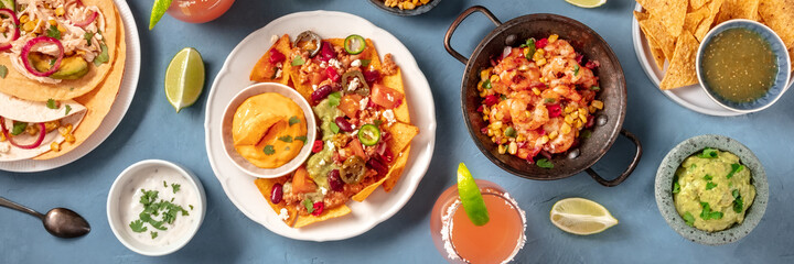 Mexican food, a flat lay overhead panorama on a blue background. Nachos, tacos, Paloma cocktails, guacamole, and various dips