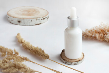 A white cosmetic bottle stands on a wooden stand on a white table, next to dried flowers. Serum, spa treatments, natural cosmetics, eco-friendly materials. Light colors. Copy space.