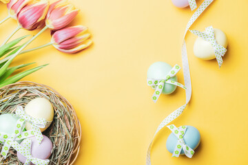 Easter holiday. Colourful egg with tape ribbon, spring tulips, feathers on pastel yellow background in Happy Easter decoration. Spring holiday top view with copy space.
