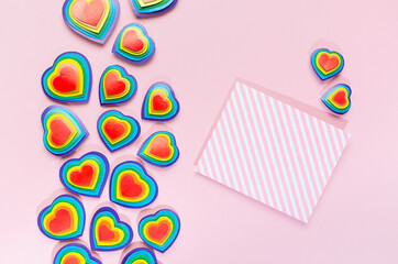Valentine lgbt festive background - rainbow bright hearts with blank striped paper for text on pastel pink backdrop, top view.