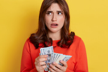 Puzzled adorable european female wearing orange sweater standing isolated over yellow background with opened mouth and looking away, holding cash in hands and think how to spend.