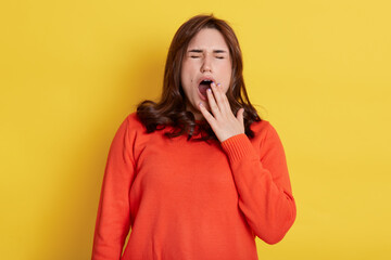 Beautiful woman yawns isolated over yellow background, wearing casual orange jumper, has dark hair, covering her mouth with palm, keeps eyes closed, feels sleepy.