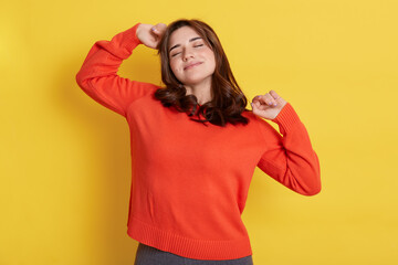 Portrait of young woman stretching and yawning against yellow background, keeps eyes closed, feels sleepy, wearing orange sweater, being lazy, just wake up.