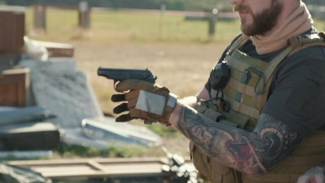 Handheld tilting-up shot of brutal tattooed military man in khaki vest loading handgun practicing aiming