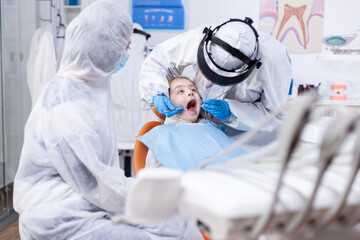 Little girl with mouth open getting caries treatment from pediatric dentist dressed in ppe suit. Dentist in coronavirus suit using curved mirror during teeth examination of child.