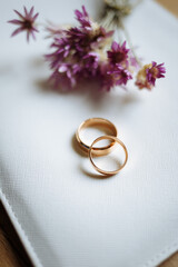 Two golden wedding rings and pink flowers on white background.