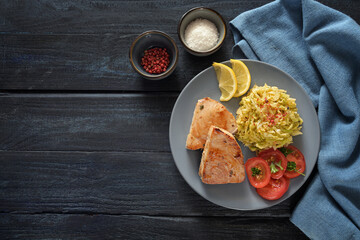 Fried tuna steaks, savoy cabbage, tomatoes and lemon on a blue plate and a dark rustic wooden table with napkin and spices, copy space, high angle view from directly above