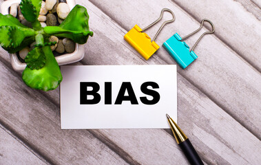 On a wooden background, a white card with the text BIAS, yellow and green paper clips and a plant...