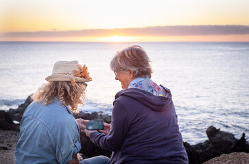 Two women sitting admiring the sunset over the sea while using their mobile phone. An elderly person and a curly middle-aged people enjoy freedom and holiday