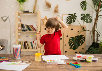 happy little boy draws with a brush and paints on white paper on a wooden desk with stationery. Creative development of the child