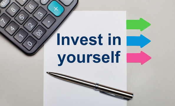 On a light gray background, a calculator, a white sheet with the text INVEST IN YOURSELF, a pen and bright multicolored stickers. View from above