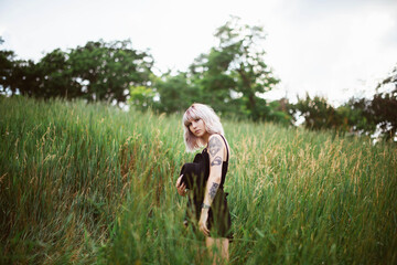 Young stylish girl with blonde hair standing in the green field in black dress, holding black hat. Portrait shot