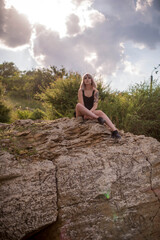 Young stylish blonde woman with tattoos in black dress sitting on a big rock outdoors on sunny day