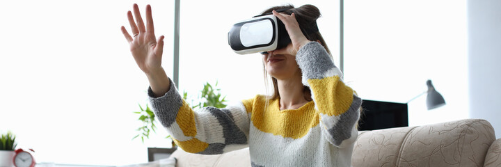 Portrait of young woman wearing virtual reality glasses and travel in different world. Beautiful female sitting on comfy couch in living room. Innovative technologies and entertainment concept