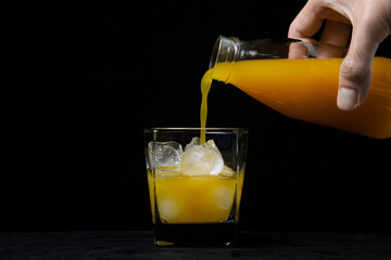 The juice is poured into a glass with ice on a black background. A male chicken pours juice from a bottle. Natural mango juice