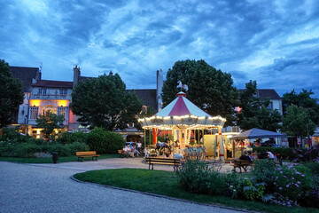 Scene of an evening in Beaune (France), representing an illuminated carousel and some people sitting on the bench around it