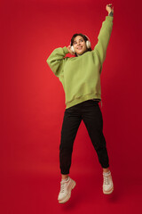 Dancing in headphones. Caucasian woman's portrait isolated on red background with copyspace. Beautiful female model in green hoodie. Concept of human emotions, facial expression, sales, ad, fashion.