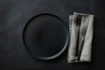 Empty black plate with black cutlery and gray linen napkin on black stone table. Top view.