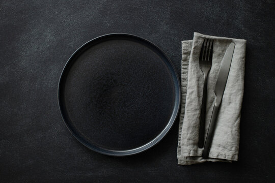 Empty Black Plate With Black Cutlery And Gray Linen Napkin On Black Stone Table. Top View.