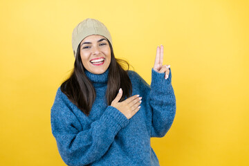 Young beautiful woman wearing blue casual sweater and wool hat smiling swearing with hand on chest and fingers up, making a loyalty promise oath