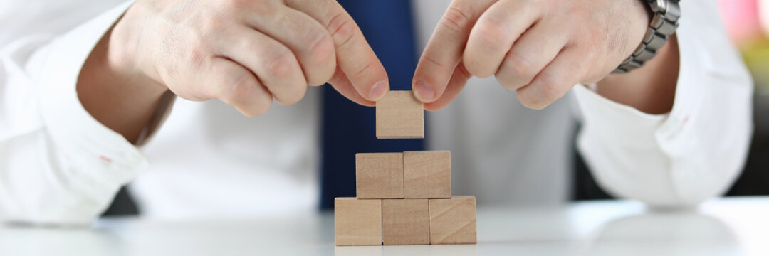 Close-up Of Businessman Putting Wooden Block On Top Of Pyramid. Thinking About Next Step. Planning Business Growth. Worker In Luxury Suit. Leadership And Smart Decision Concept