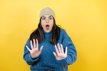 Young beautiful woman wearing blue casual sweater and wool hat afraid and terrified with fear expression stop gesture with hands, shouting in shock. Panic concept.