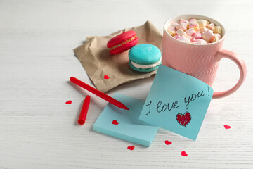 Note with phrase I Love You near pink cup of hot drink and macarons on white wooden table