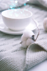 Fototapeta na wymiar Large white ceramic cup on a saucer and a sprig of cotton on a knitted nude blanket. Cozy home background with blur. Still life from home interior