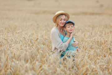 Mom with a straw hat and blue dress hugs her son in a cap standing in a wheat field. Summer holidays in the village