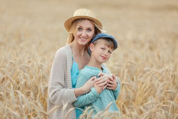 Naklejka na ściany i meble A portrait of a mother in a straw hat and blue dress hugs her son in a cap standing in a wheat bread field. Summer holidays in the village
